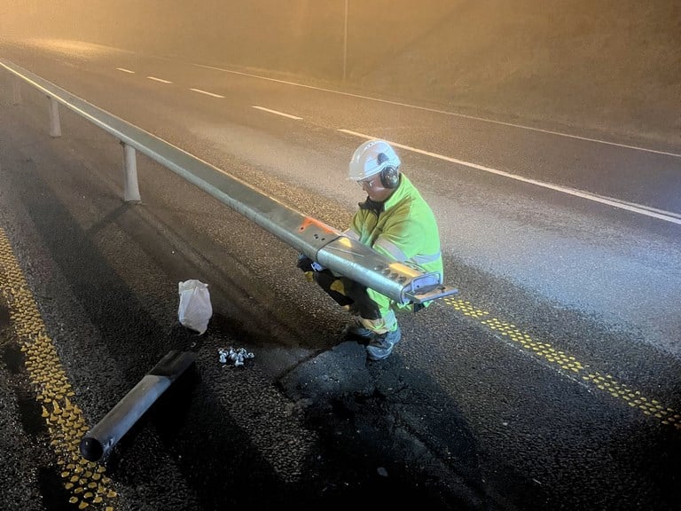 Mann med gul vernejakke og hjelm sitter på huk ved midtrekkverket og monterer et element på midrekkverket. 