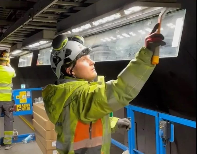 Mann med gul jakke og hjelm installerer lys i armatur i tunnel.