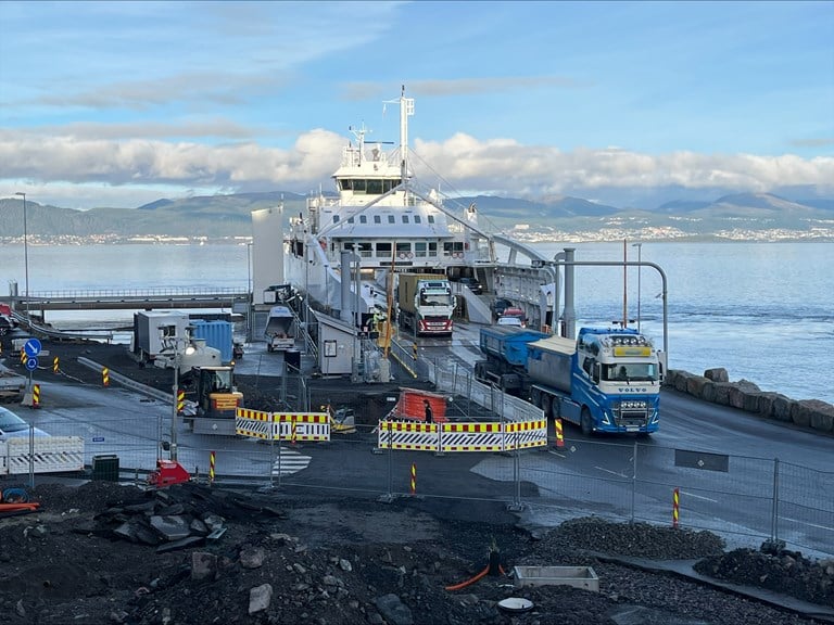 Bilete av anleggsplassen og ferjekaia, medan ferje ligg til kai og køyretøy køyrer i land.