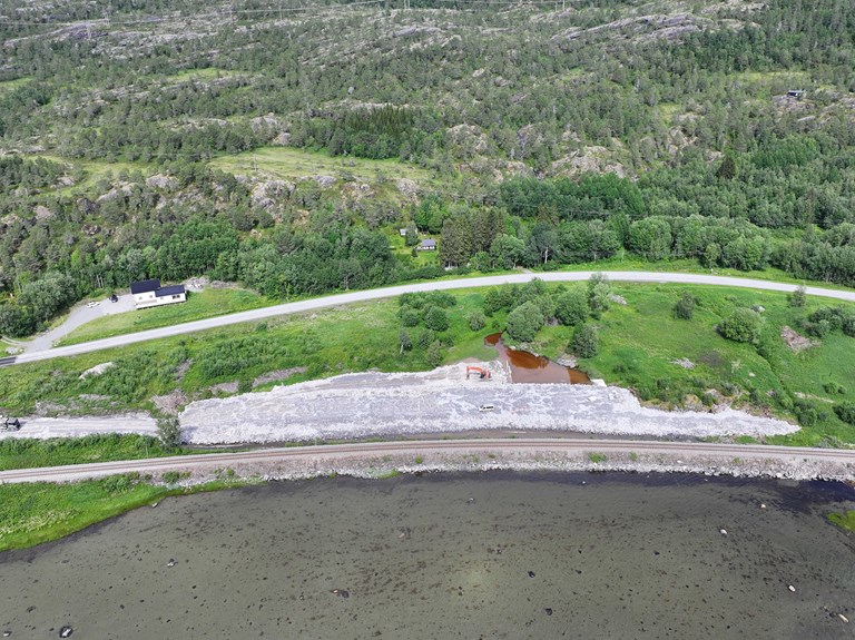 Dronebilde av steinfylling med bildeteksten: Fra den nye vegtraseen på Kistrand skal det legges stikkrenne under Nordlandsbanen. Jobben vil medføre støyende nattarbeid.