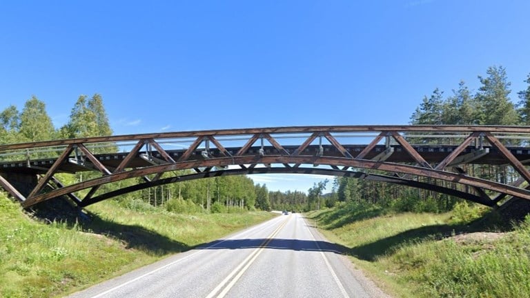 Bilde av en fagverksbru i tre som går over en trafikkert veg, under blå himmel og omkranset av grønn vegetasjon. 