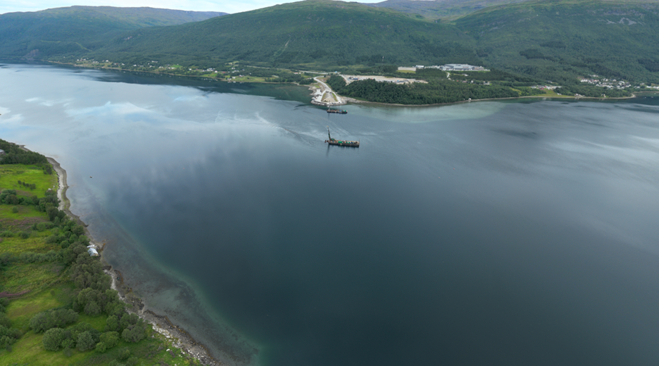 På bildet ser vi et dronefoto over Ramfjorden, hvor vi ser mot sjøfyllingen som er lagt opp i Sørbotn. I Ramfjorden ligger det to lektere som holder på med pelearbeid til pelegruppene som skal fundamentere brua. 